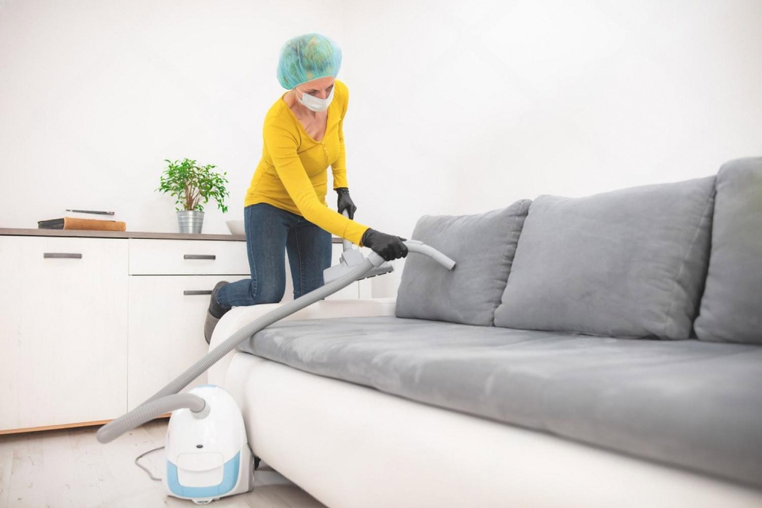 Cleaner in a mask vacuuming a grey fabric sofa in a Dubai apartment