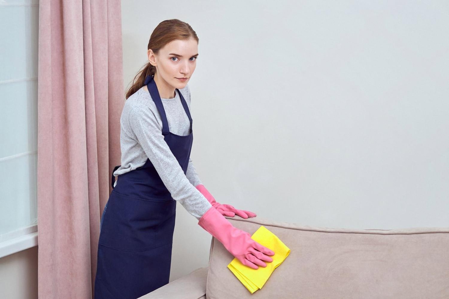 Housekeeper in pink gloves wiping the back of a beige sofa near a window